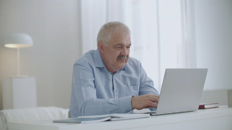 Un-Anciano-De-Pelo-Gris-Está-Trabajando-Remotamente-Desde-Casa-Comunicándose-Mediante-Chat-Escribiendo-Mensajes-En-Un-Portátil-Con-Conexión-A-Internet